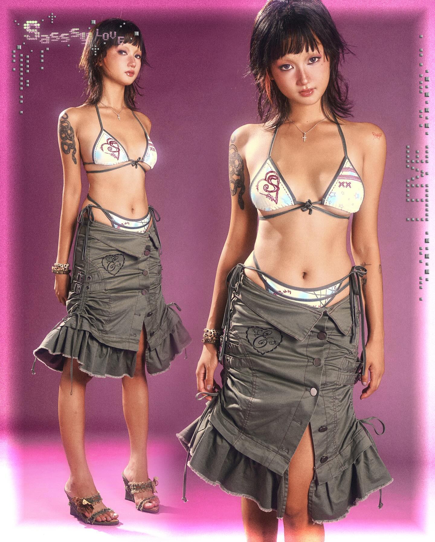 the image features two identical young women, likely twins, showcasing a matching outfit. they are both wearing a khakigreen, ruffled miniskirt with a drawstring closure and a lightcolored bikini top with a heart design.  both models sport tattoos and are wearing sandals with heels. the background is a vibrant pink, and the overall style is suggestive of high fashion, summer, and a youthful aesthetic. the image has a playful and slightly edgy feel due to the styling and pose.  the words "sassysolove" are visible, potentially indicating a brand or label.