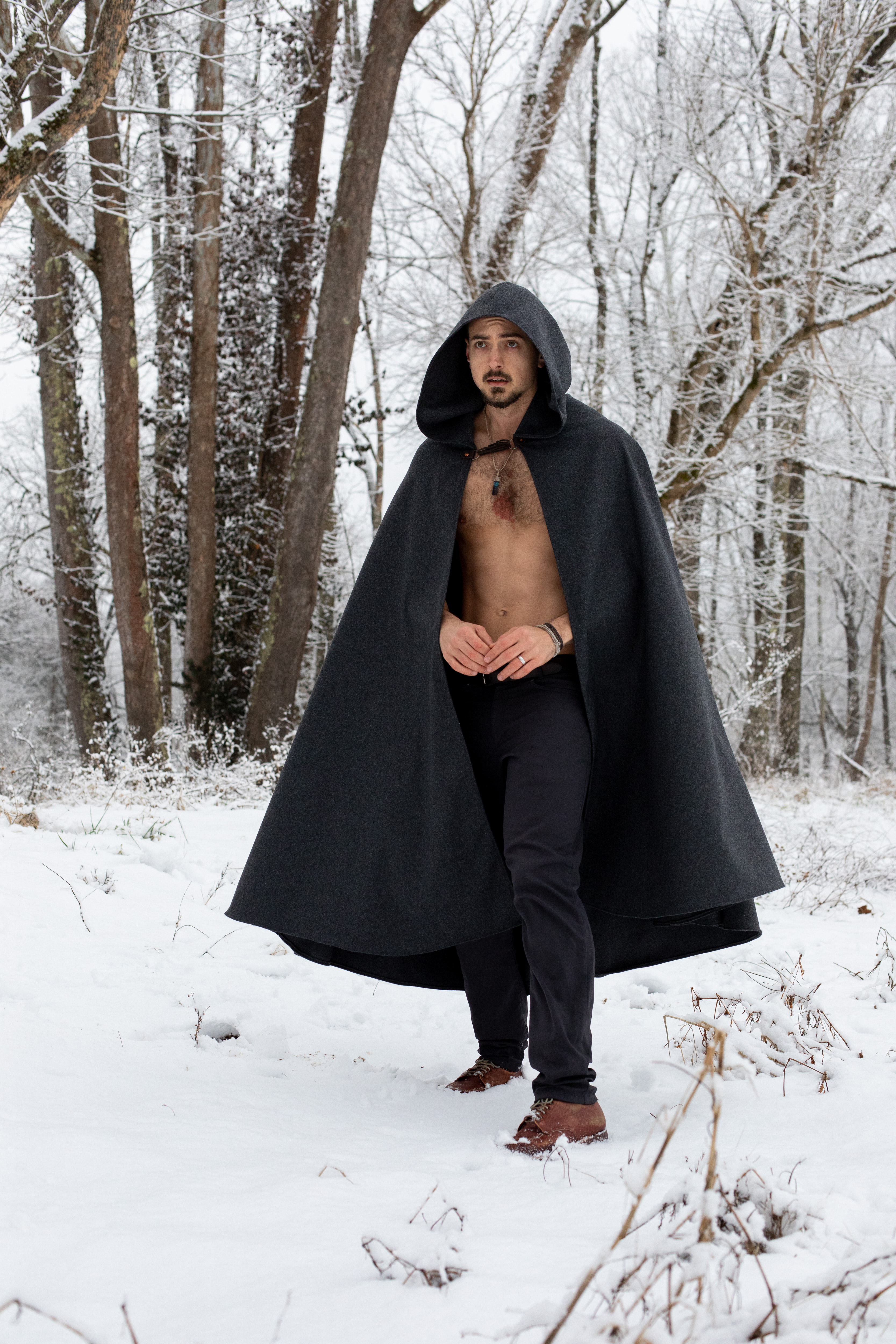 a man with a bare chest and a black cloak stands in the snowy woods. he is wearing dark pants and brown shoes and has a silver chain around his neck. the trees and ground are covered in snow, and the sky is overcast.
