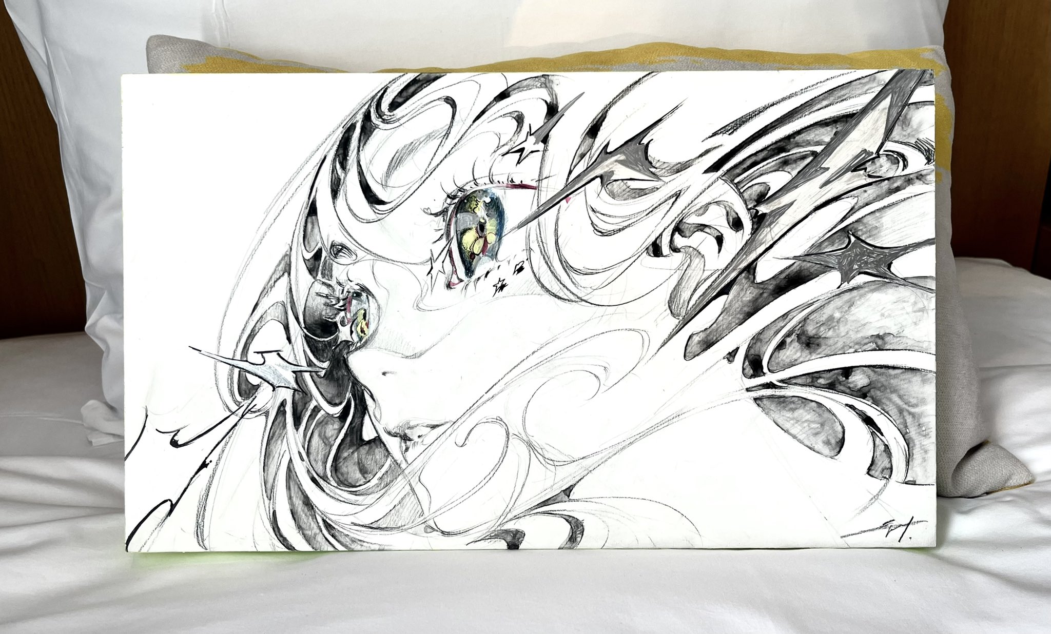 a pen and ink sketch predominantly in black and white, resting on a bed with white linens and a yellow pillow. the artwork features an abstract portrait of a woman, her eye emphasized amidst a whirlwind of swirling lines. the style suggests a blend of realism and abstract expressionism.
