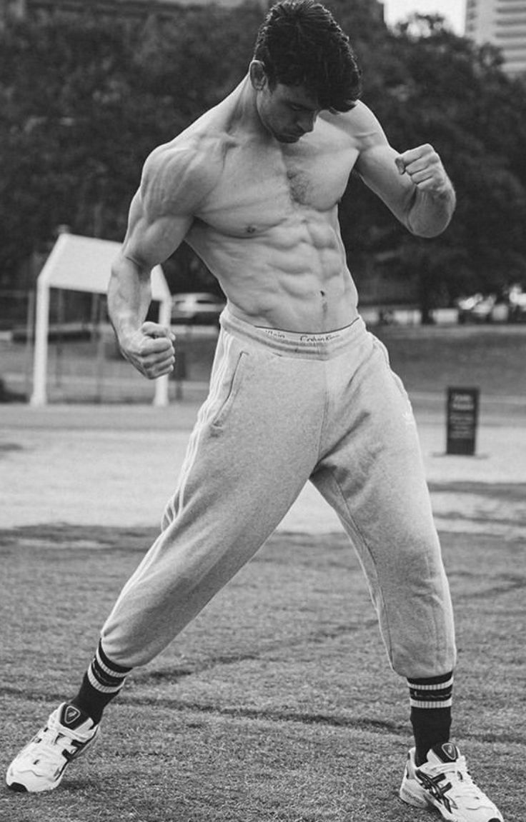 a shirtless man with a muscular physique, wearing grey sweatpants and athletic shoes, is shown standing outside in a grassy field. he has one arm extended and his fist clenched.