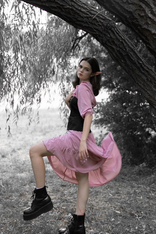 a woman dressed as an elf stands in front of a tree. she wears a pink dress and black boots, and has pointed ears. the background is gray and the woman is in color.