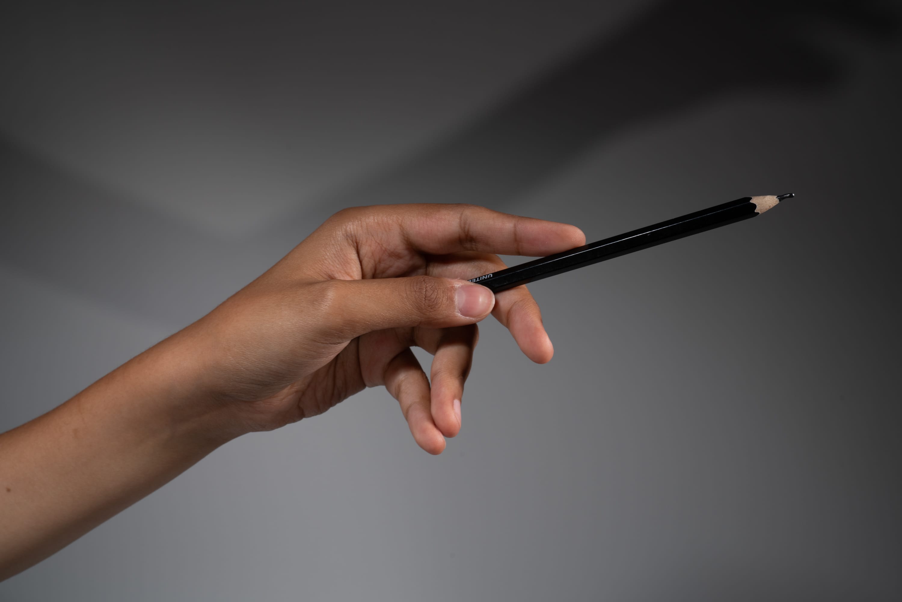 a hand is holding a black pencil against a gray background. the hand is pointing the pencil to the right side of the image. the pencil is casting a shadow on the wall.