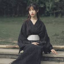 a young woman wearing a black kimonostyle dress is sitting on stone steps in a forested area. she has long dark hair and is looking slightly to the left of the camera. the lighting is soft and natural, creating a serene and contemplative mood.