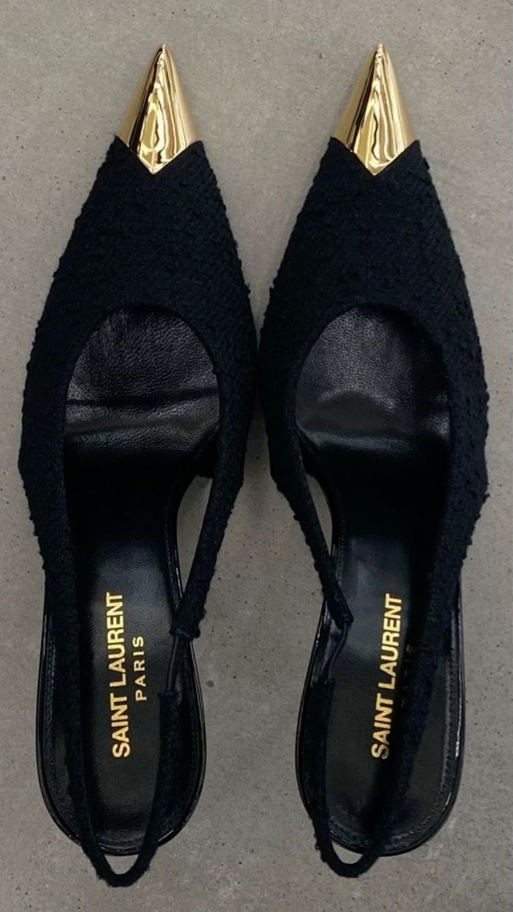 a pair of black saint laurent slingback heels with gold pointy toes are shown from above against a grey background.  the shoes have a textured fabric upper and the "saint laurent paris" logo is visible inside.  the heels are positioned slightly apart, with their toes pointing upwards.