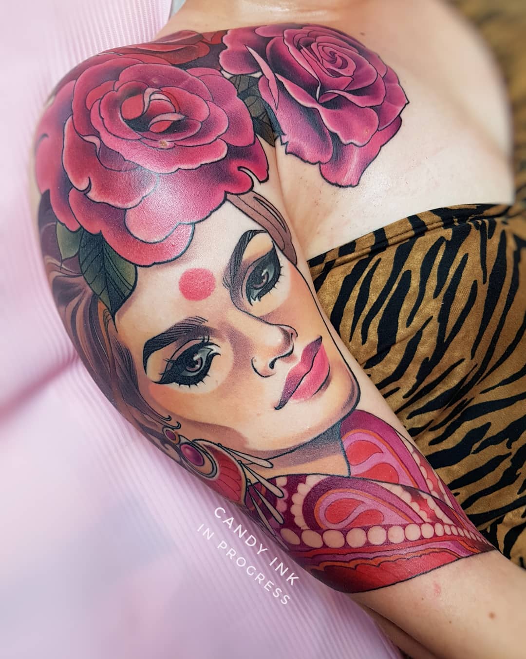 the image shows a tattoo artist's perspective of a colorful, inprogress tattoo on a client's upper arm. the tattoo features a stylized portrait of a woman with dark hair, red lipstick, and a prominent bindi, adorned with large, vibrant pink roses above her head.  she is wearing traditionallooking clothing with intricate patterns in shades of red and pink. the tattoo style is bold and graphic with heavy black outlines. the client is wearing a zebraprint top.