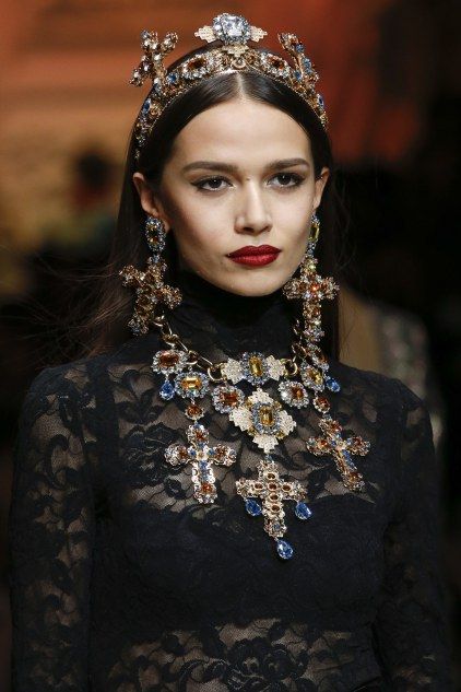 a closeup shot of a fashion model on a runway, wearing a stunning dolce & gabbana ensemble. the model is adorned with an elaborate crown and matching necklace and earrings, all featuring cross motifs and an abundance of crystals and gemstones. the black lace top adds a touch of gothic elegance. the model's dark hair and striking red lipstick perfectly complement the opulent jewelry. the overall image exudes luxury and high fashion.