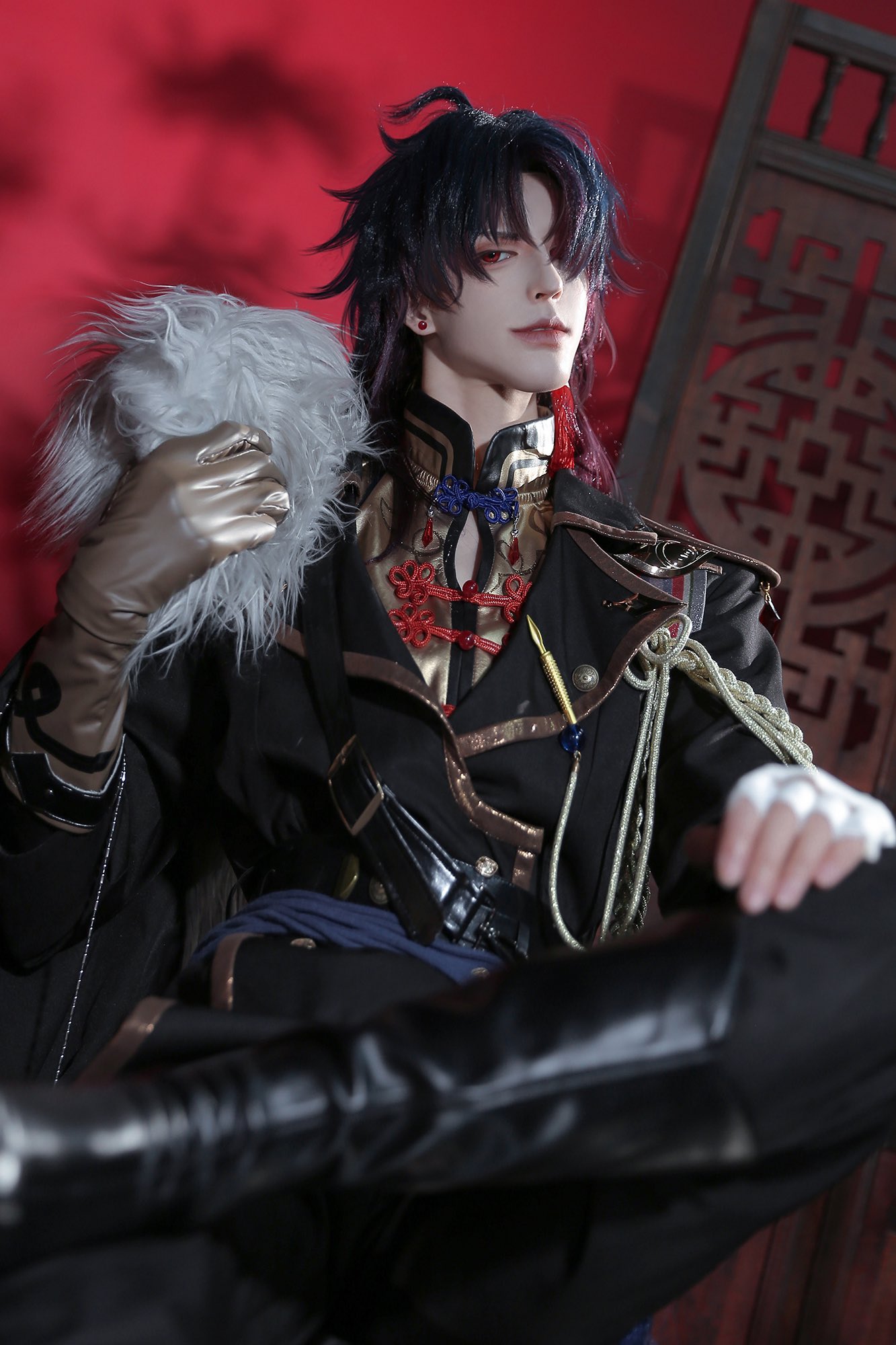 a young man dressed in a chineseinspired cosplay outfit. he is sitting in a chair with a serious expression on his face. the background is a red wall with intricate chinese patterns. he is wearing a black jacket with gold trim, a white and black shirt with red accents, black pants, silver and black gloves and black boots. he has long black hair and is holding a white fluffy item. the light hits the man's face, giving him an almost ghostly appearance.