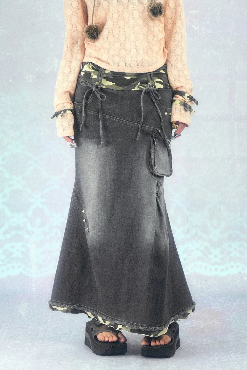 a fulllength image showcasing a model wearing a long, grey denim skirt. the skirt features a distressed look, an asymmetrical design, multiple pockets, and a drawstring waist with a camouflage fabric accent underneath.  it is layered over a lighter colored fabric. the model also wears platform sandals and a light peachcolored longsleeved top. the background is a soft, light blue/purple. the overall style is a mix of boho and grunge influences.