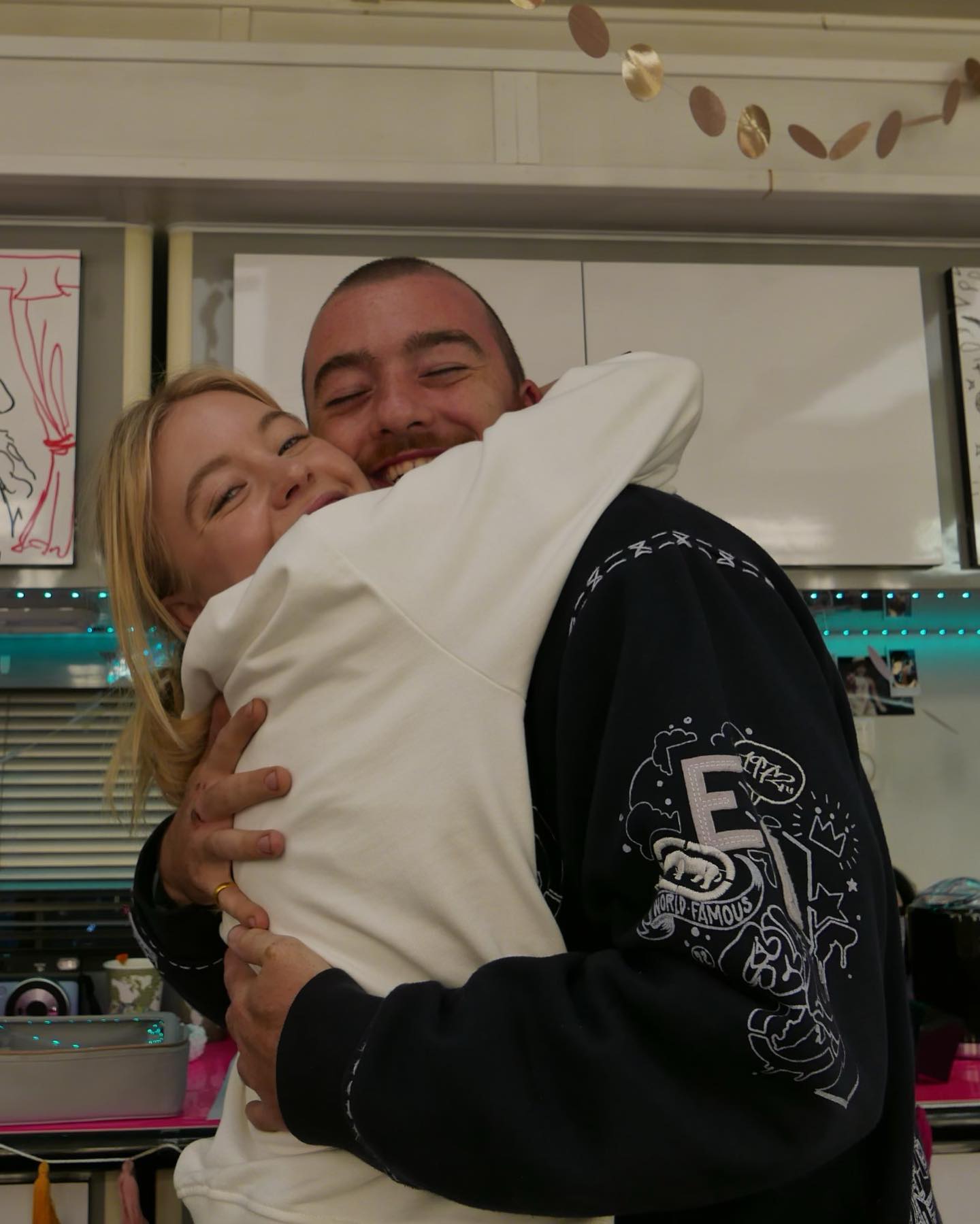 the image shows a young man and woman in a warm embrace. the woman has blonde hair and is wearing a white sweatshirt. the man has short dark hair and is wearing a darkcolored jacket with graphic design on the sleeve.  they appear happy and affectionate, with the woman's arms wrapped around the man's neck and the man's arms around her waist. the background suggests an indoor kitchen setting, with some kitchen appliances and cupboards partially visible. the overall mood of the picture is positive and conveys a sense of closeness and connection between the two individuals.