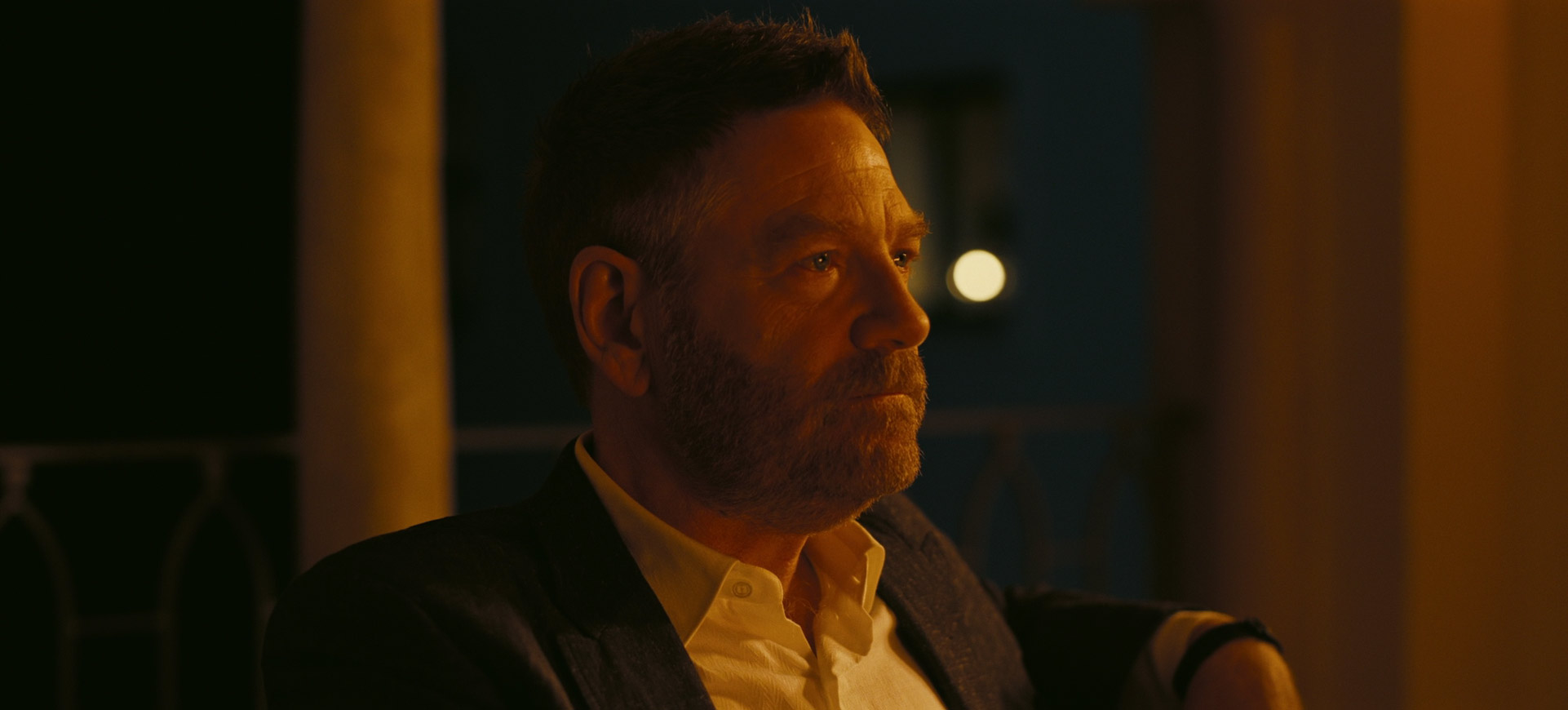 a man with a beard sits in a dark room, looking thoughtfully towards the right side of the frame. he's wearing a white buttondown shirt and a jacket. the light illuminates his face and upper body, but the rest of the scene is mostly dark. he appears to be in deep thought.