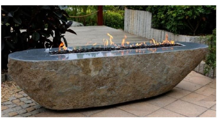 a stone fire pit is shown outdoors. the fire pit is large and rounded, with a stone base and a rectangular metal firebox filled with black lava rocks. flames are visible in the firebox. the fire pit is situated in a patio or garden setting, with landscaping and hardscaping in the background.
