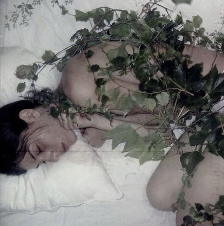 the image shows a woman sleeping in bed, surrounded by vines and leaves. she is lying on a white pillow and white bedding. the vines are green and lush, covering part of her body. the scene evokes a feeling of nature intertwined with human rest.