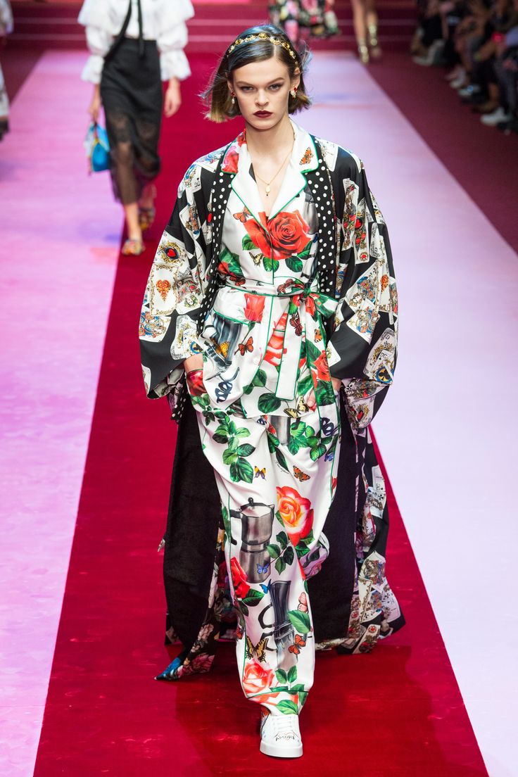 a model walks the dolce & gabbana spring/summer 2017 runway wearing a stunning pajamainspired pantsuit. the ensemble features a vibrant floral print with roses, coffee pots, and playing cards, set against a white background.  a black kimonostyle robe with similar print details adds a layer of sophistication. the model's hair is styled in a bob with a gold headband, and she wears white sneakers. the runway is a deep red carpet, and other models can be seen in the background. the overall image is one of high fashion, elegance, and luxury.
