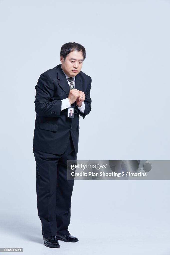 a man in a suit and tie is standing in a studio. he has his hands clasped in front of him and looks nervous or anxious. he is wearing a badge around his neck. he is wearing black dress shoes.