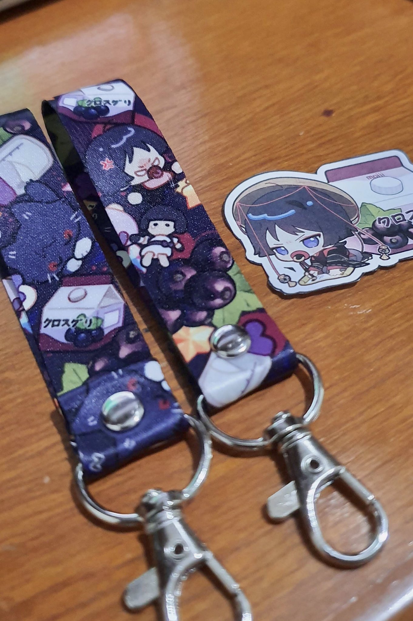 two anime lanyards with blackcurrant and milk carton designs and one anime character keychain. the lanyards have silver clips at the ends and the keychain is made of plastic with silver metal loop. the background is a wooden surface.
