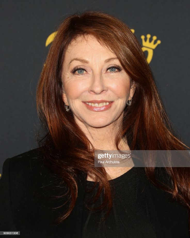 a woman with long red hair smiles at the camera. she is wearing a black top and has a dark background behind her.