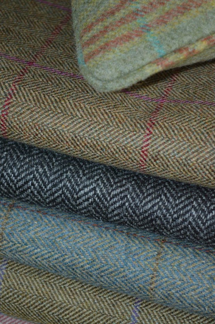 a stack of four different tweed fabrics with herringbone patterns and varying colors of brown, black, and blue. the topmost fabric is light green and brown with a plaid pattern. the bottom fabric is a natural brown tweed. the image shows the various textures and patterns of tweed fabrics.