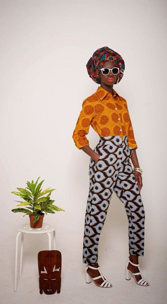a fulllength studio shot of a darkskinned woman modeling a stylish african outfit. she is wearing a vibrant mustard yellow shirt with a circular pattern, paired with light blue and brown geometric print pants.  her head is wrapped in a colorful ankara print headwrap, and she accessorizes with white sunglasses and white highheeled sandals. she is standing next to a small white stool upon which a small potted plant and a wooden mask are placed. the background is plain white, making the model and her outfit the focal point of the image.  the overall style is bold, modern, and incorporates elements of traditional african design.