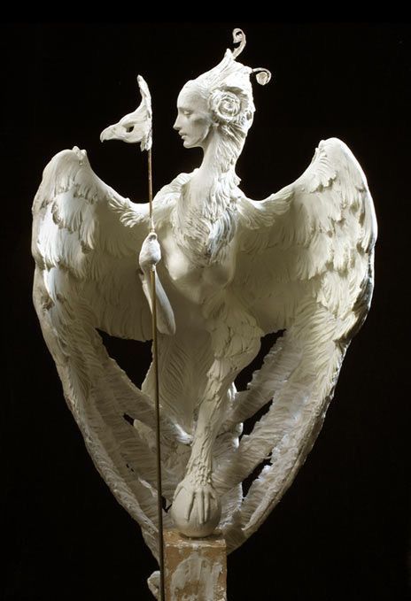 a detailed sculpture of a sphinx is shown against a black background. the sphinx is white, giving it a pristine, ethereal quality. it has the head and torso of a woman, with delicate facial features and an ornate hairstyle featuring a roselike ornament. the creature possesses large, feathered wings that spread out dramatically behind it. in its hand, it holds a stick with a bird mask. the sphinx stands atop a base, blending human and avian elements in a striking mythological portrayal.