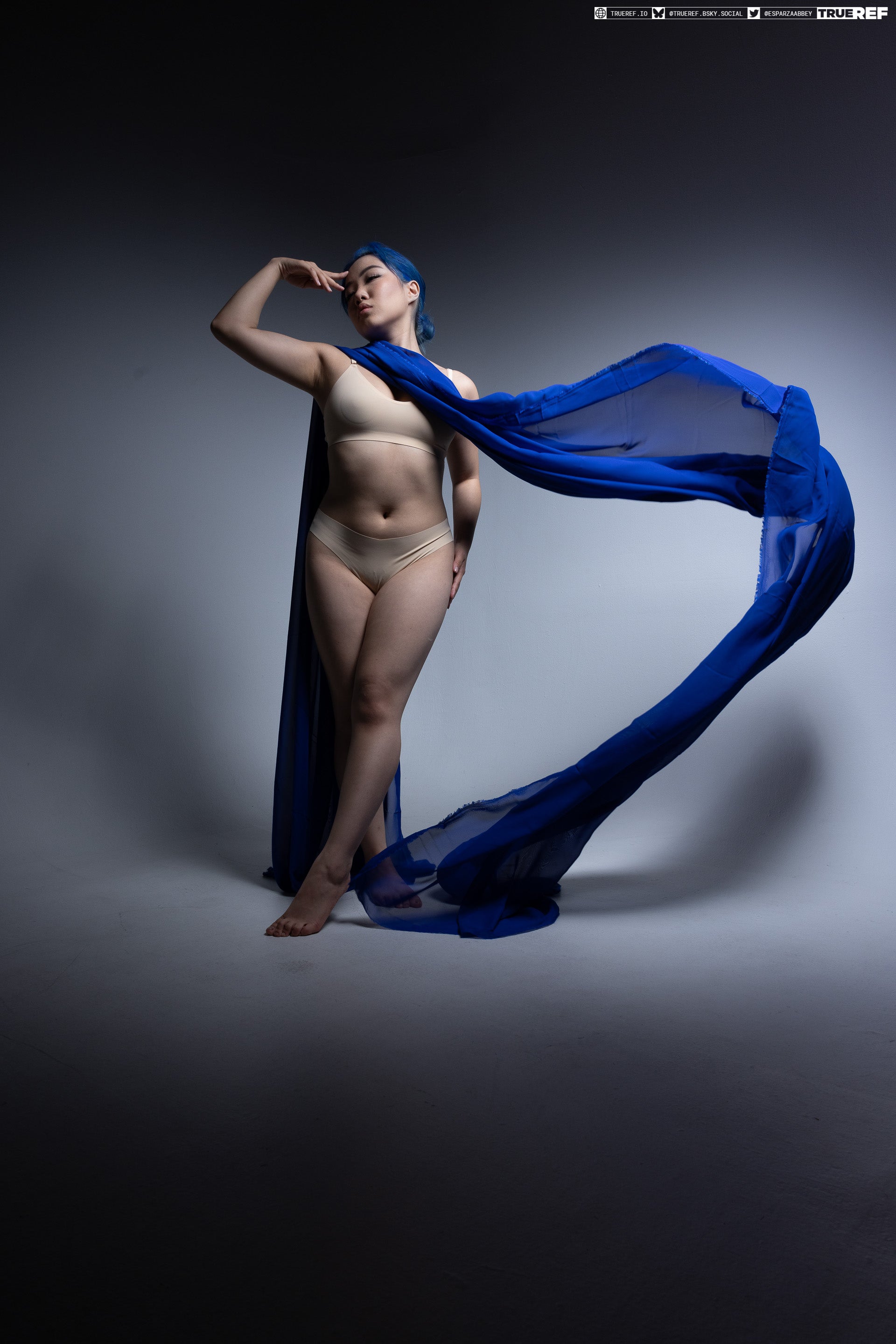 a full shot of a woman with blue hair, wearing lingerie and a blue cape, posing in a studio setting. she is looking away with her hand on her forehead. the lighting is dramatic, with a gradient background.