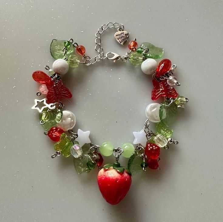 the image shows a colorful charm bracelet with a strawberry theme. the bracelet features a variety of beads in red, green, and white, along with charms such as stars, leaves, and a larger strawberry charm hanging from the center. there is also a silver "made with love" charm attached to the chain. the bracelet is made up of various beads and charms.