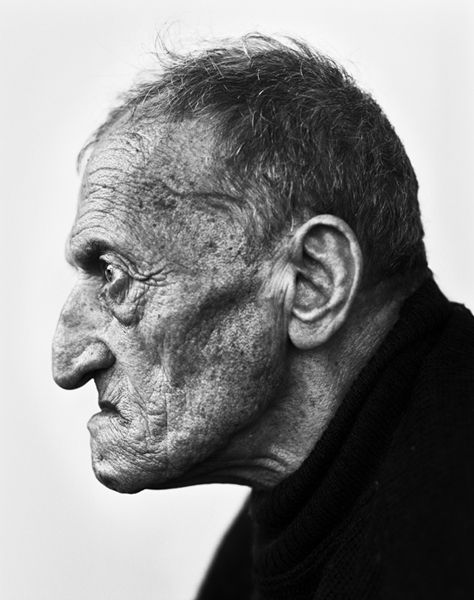 a black and white profile portrait of an elderly man with numerous wrinkles, capturing the essence of aging. he has a large nose, a pronounced ear, and sparse hair. he's wearing a dark, highnecked garment. the background is a plain white, emphasizing the man's features.