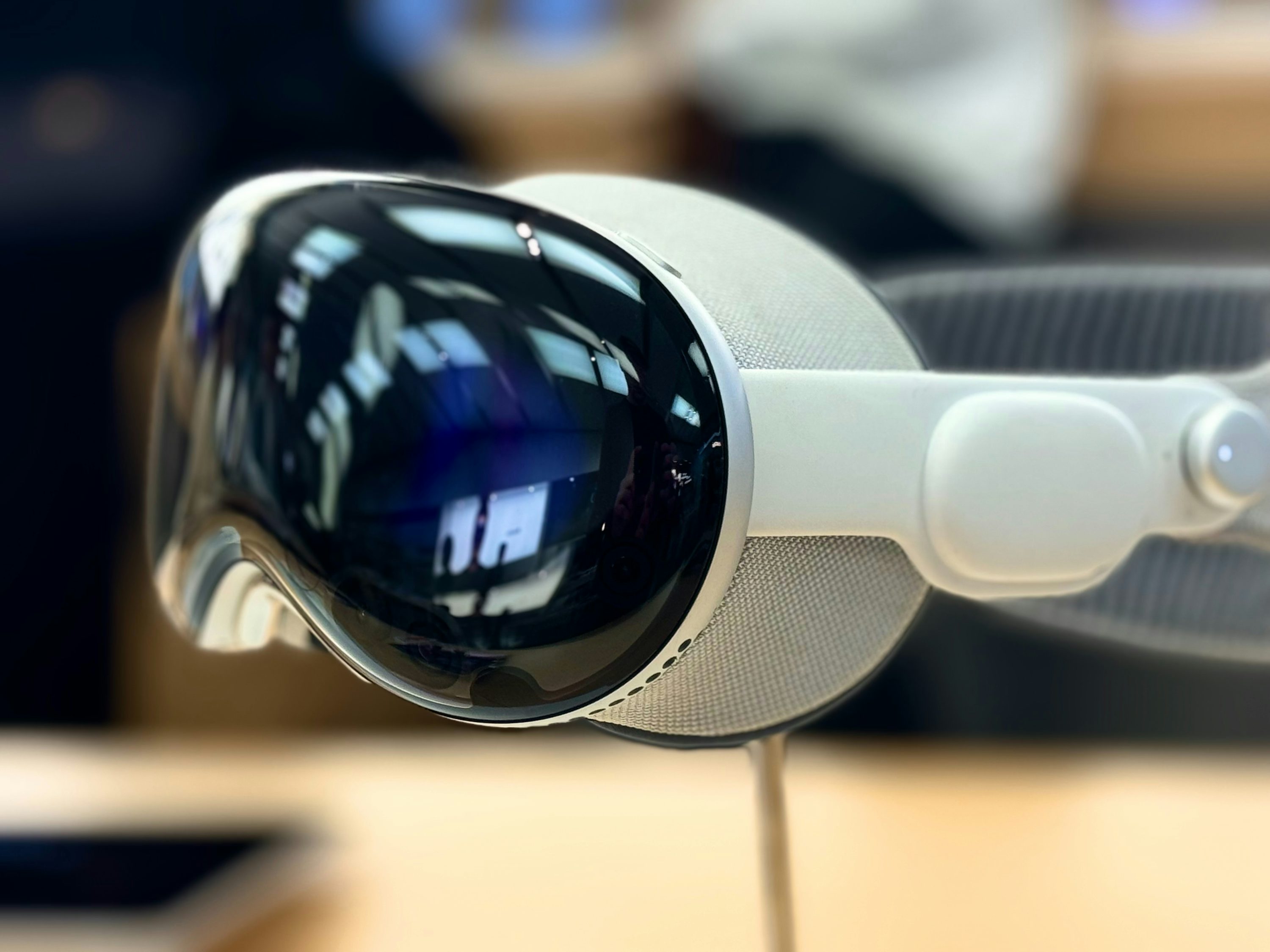 closeup shot of the apple vision pro vr headset sitting on a surface. the headset has a glossy black front visor with reflections, a white frame, and a gray fabriccovered head strap.