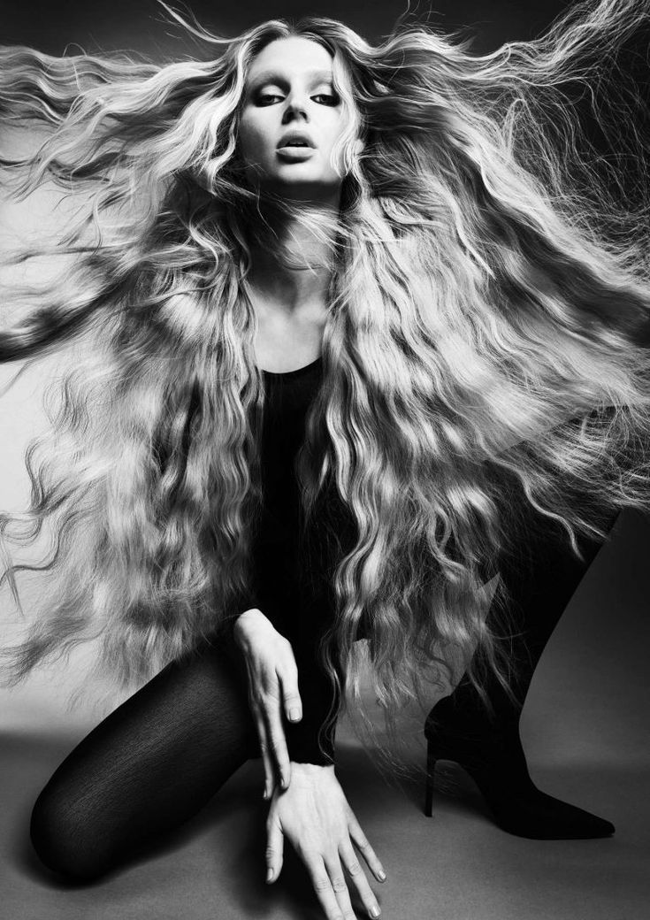 the image is a striking black and white fashion photograph of a female model with extraordinarily long, wavy blonde hair.  her hair is the central focus, dynamically flowing around her. she is kneeling, wearing a black bodysuit and tights, and black heeled boots. her pose is dramatic and slightly contorted, emphasizing the length and movement of her hair. the lighting is dramatic and creates strong contrasts and shadows, highlighting the texture of her hair and clothes. the overall mood is elegant, artistic, and somewhat mysterious.