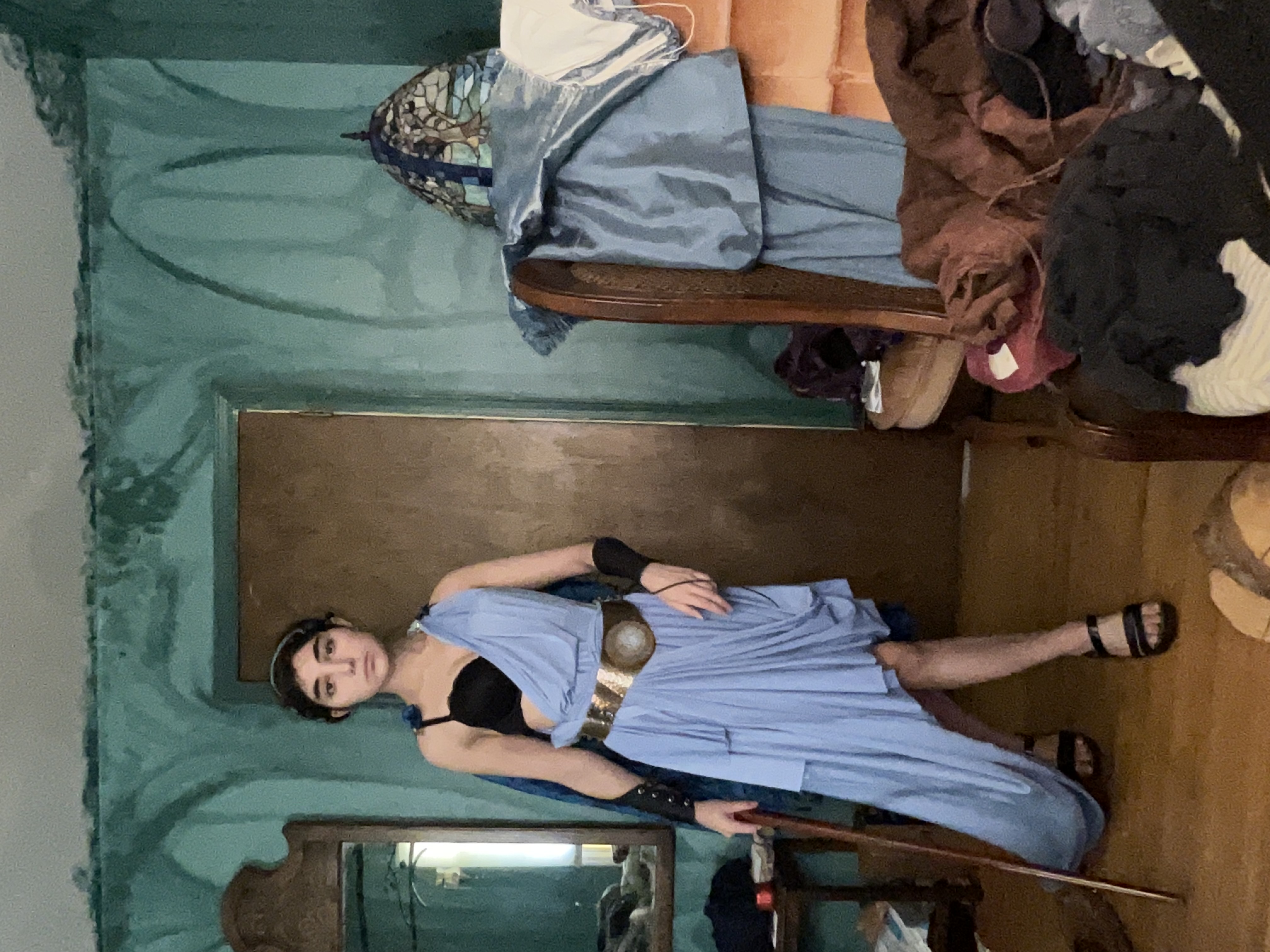 a young woman is posing in a cosplay costume resembling an ancient greek goddess. she is wearing a flowing light blue asymmetrical dress, a gold belt, dark sandals, and a headband. she holds a long wooden staff. the setting is a messy bedroom with a vintage chair and a stained glass lamp visible in the background.  the walls are painted a teal color with a textured, somewhat artistic design.