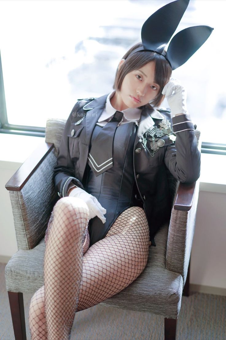 a person dressed in a bunny girl military uniform is sitting in a chair. the person has short hair and is wearing bunny ears, a military uniform, fishnets, and gloves. the person is sitting in a gray chair indoors.