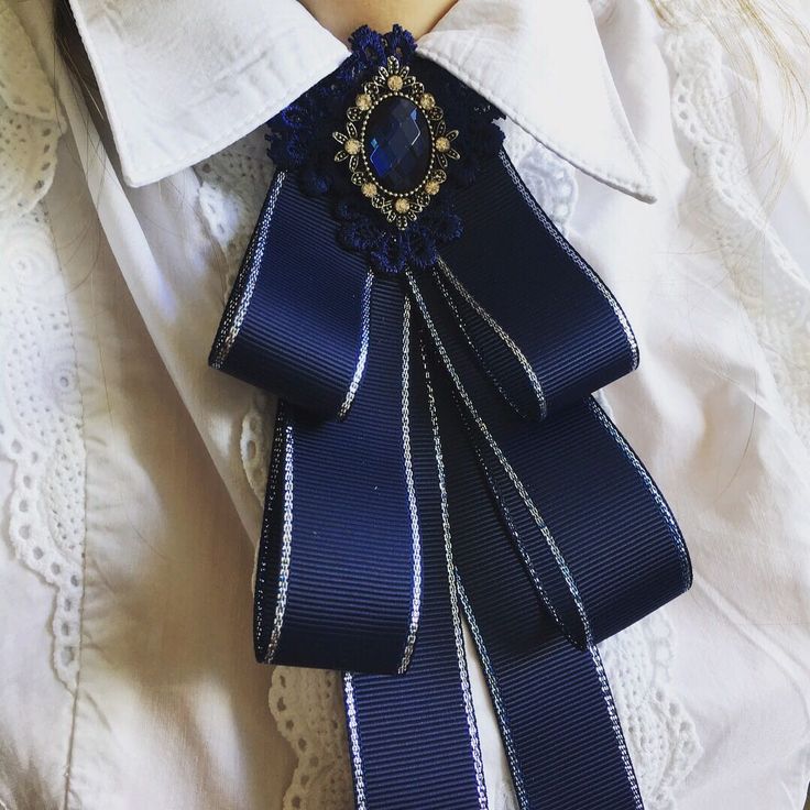 the image shows a closeup view of a navy blue bow tie with a silver trim worn on a white lacetrimmed shirt. the bow tie is embellished with a large, ornate brooch featuring a dark blue gemstone surrounded by goldtoned metal and delicate lacework.  the fabric of the bow tie appears to be a stiff, structured ribbon. the overall style is elegant and reminiscent of vintage or victorian fashion. the focus is on the intricate detail of the bow tie and its embellishments against the backdrop of the white shirt.