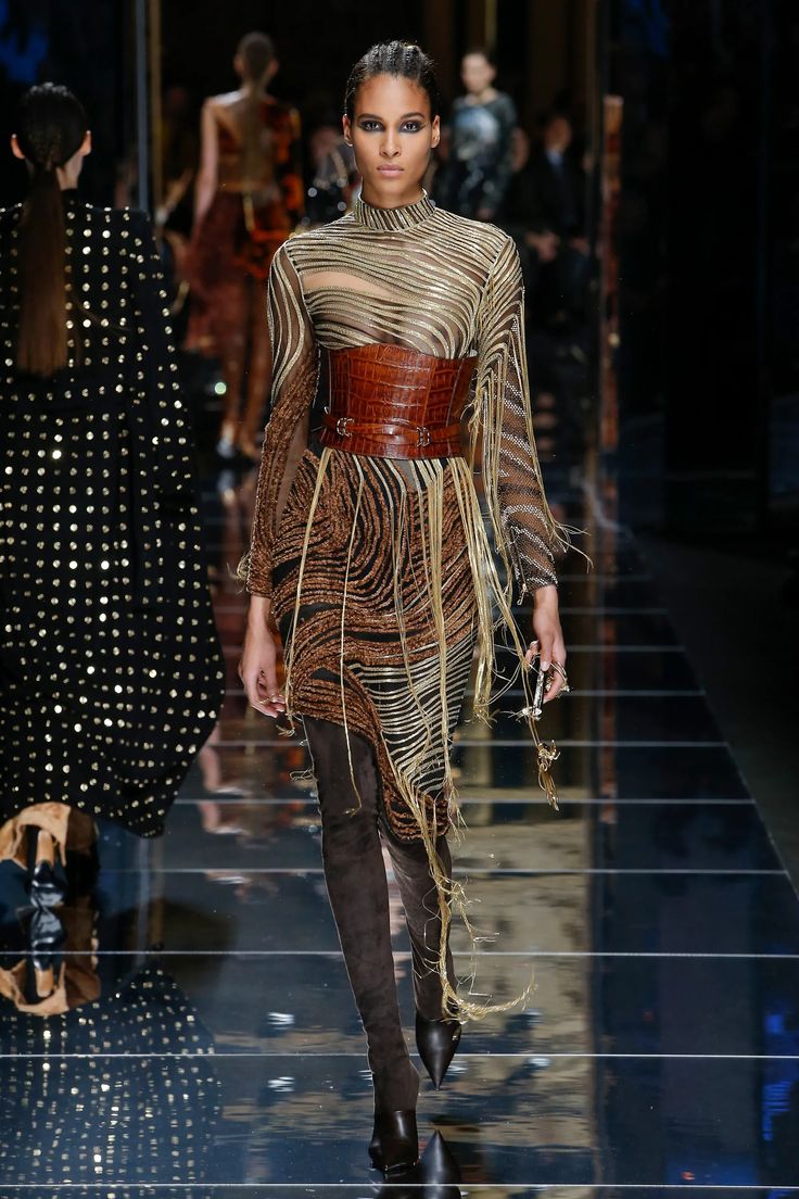 a female model walks down the runway in a brown, gold and black dress with a leather belt and thighhigh boots. her hair is pulled back and her makeup is minimal. the background is a blurred image of the runway and other models. the dress is a long, flowing design with a sheer top and a bottom that appears to be made of fringe or tassels. the belt is wide and features a gold buckle. the model's pose is confident and graceful.
