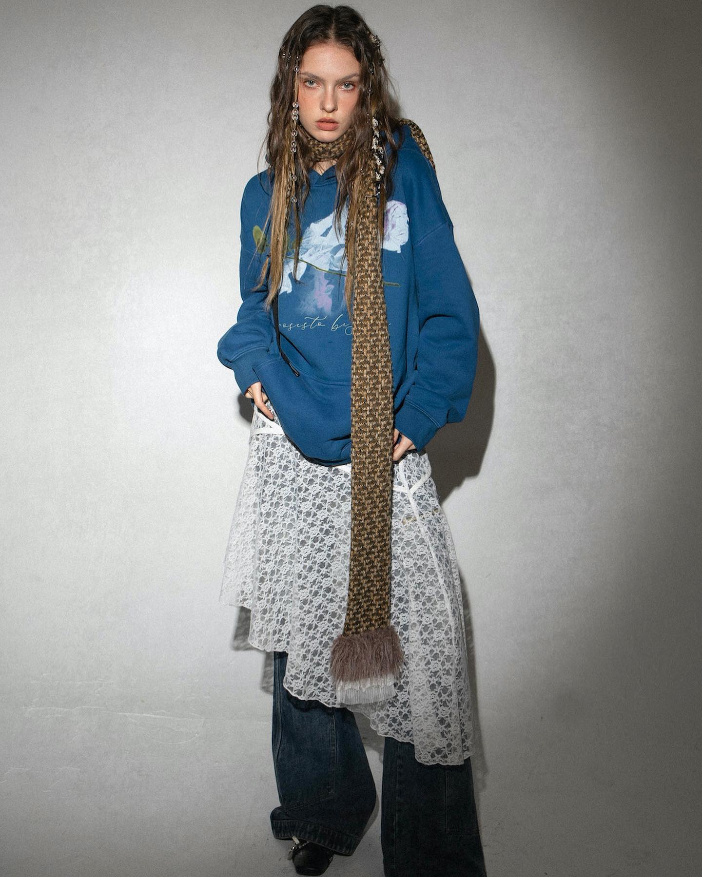 a fulllength shot of a young woman, styled in a casual yet fashionable outfit. she's wearing an oversized, dark blue hoodie featuring a faint, artistic print.  the hoodie is paired with a white asymmetrical lace skirt that falls to her midcalf, and loosefitting denim jeans are worn underneath. a long, chunky, brown and cream knitted scarf is draped around her neck, complementing the overall relaxed aesthetic. her long brown hair is loose, and she appears to be posing in a studio setting against a plain grey backdrop.  the style is distinctly bohochic with a contemporary twist.