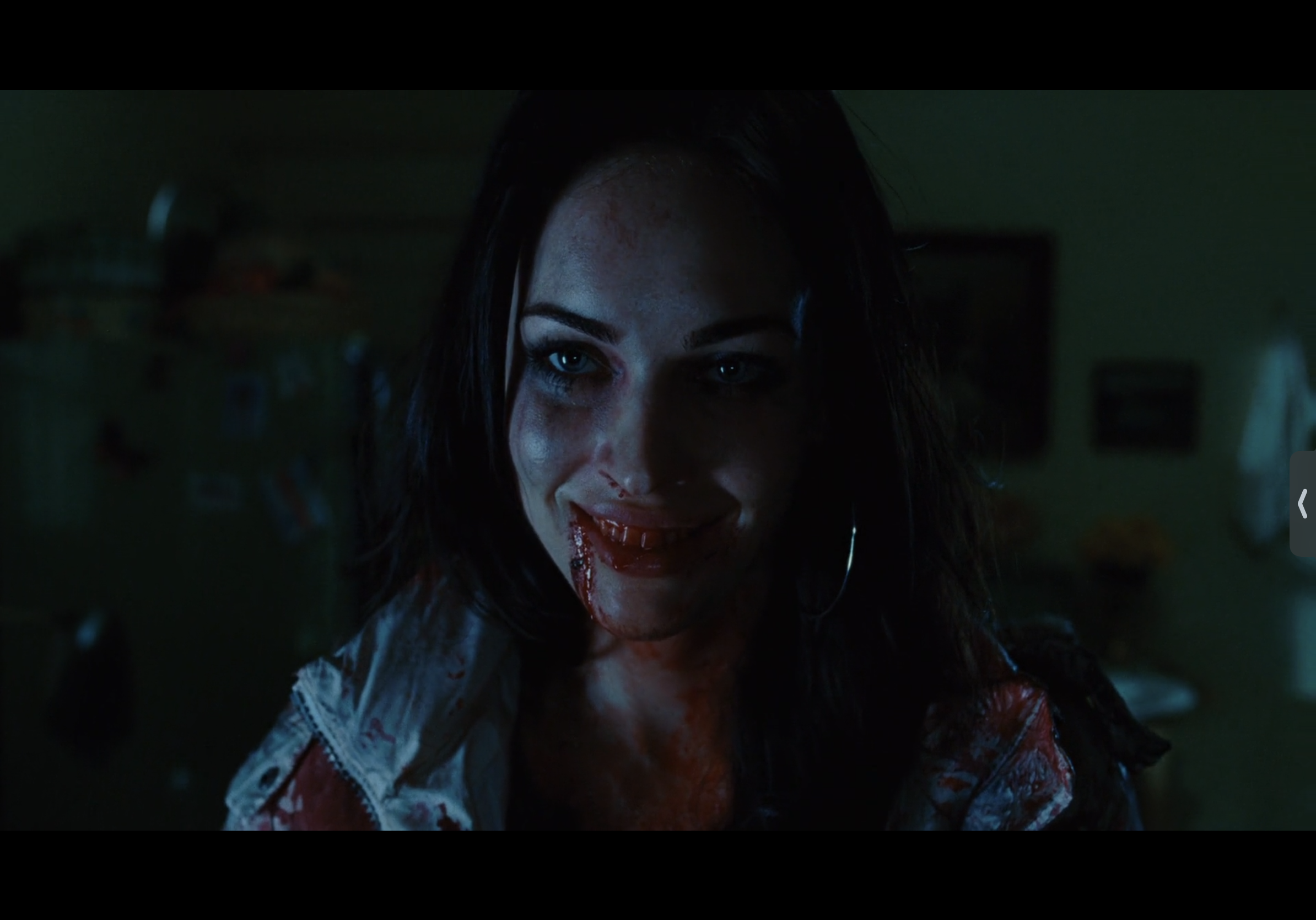 a young woman with long dark hair is covered in blood, she smiles creepily with blood around her mouth, she is standing in a dimly lit room.
