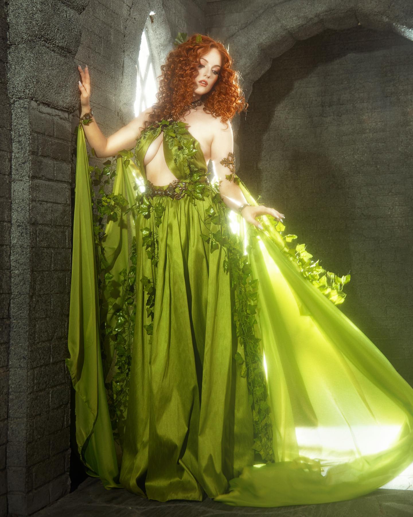a woman with long, flowing red hair stands in a stone archway. she wears a flowing green gown adorned with ivy, evoking a sense of nature and fantasy. the soft lighting and her elegant pose contribute to the overall mythical and ethereal atmosphere of the image.