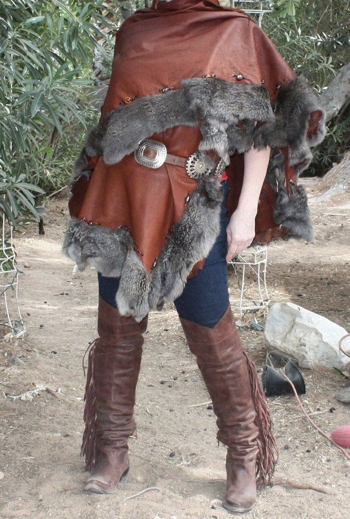 a woman is wearing a brown leather poncho trimmed with grey fur. she is wearing a belt with a silver buckle over her jeans. she is wearing brown leather boots with fringe. the photo is taken outdoors in a rural setting.