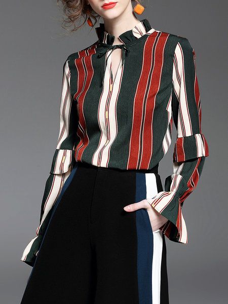 a woman with long dark hair wearing a longsleeve red and green striped shirt with ruffles around the neck and sleeves. the shirt is tucked into black pants with blue and white stripes down the sides. she has a red lip, earrings, and a neutral face expression. the background is a light gray.