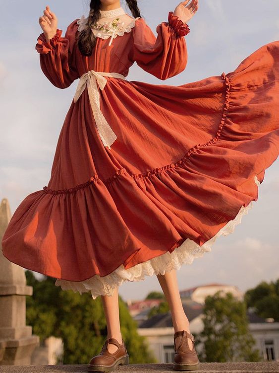 a woman in a long, flowing burnt orange dress stands on a rooftop, her arms outstretched. the dress has a lace collar, puffy sleeves, a tiered ruffle skirt, and a white lace petticoat. the golden hour sunlight casts a warm glow on the scene. the woman's pose and the dress's movement evoke a sense of freedom and joy.
