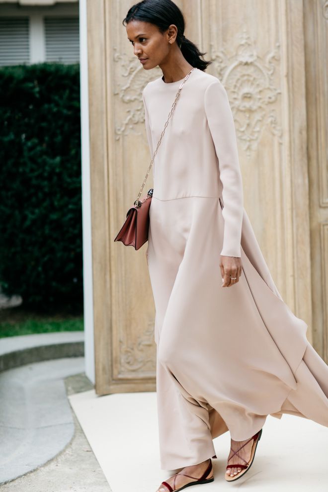 the image shows a stylish woman walking down the street. she is wearing a long, flowing light beige maxi dress with long sleeves. the dress has a simple, aline silhouette that flows elegantly.  she carries a burgundy crossbody bag and is wearing dark red velvet flat sandals with thin straps. her hair is pulled back in a simple low ponytail, and her makeup is minimal. she is walking past an ornate wooden door of an old building, and the overall setting appears to be a european city. the image's style is sophisticated and minimalist.