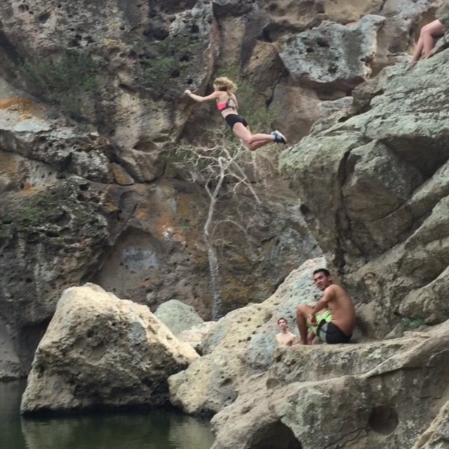 a woman is seen cliff jumping into a body of water.  she is wearing a bikini top and bottoms. below, two men are seated on rocks, watching. the surrounding environment is a rocky canyon. the water at the bottom is dark and still.  the scene is sunny and outdoors.