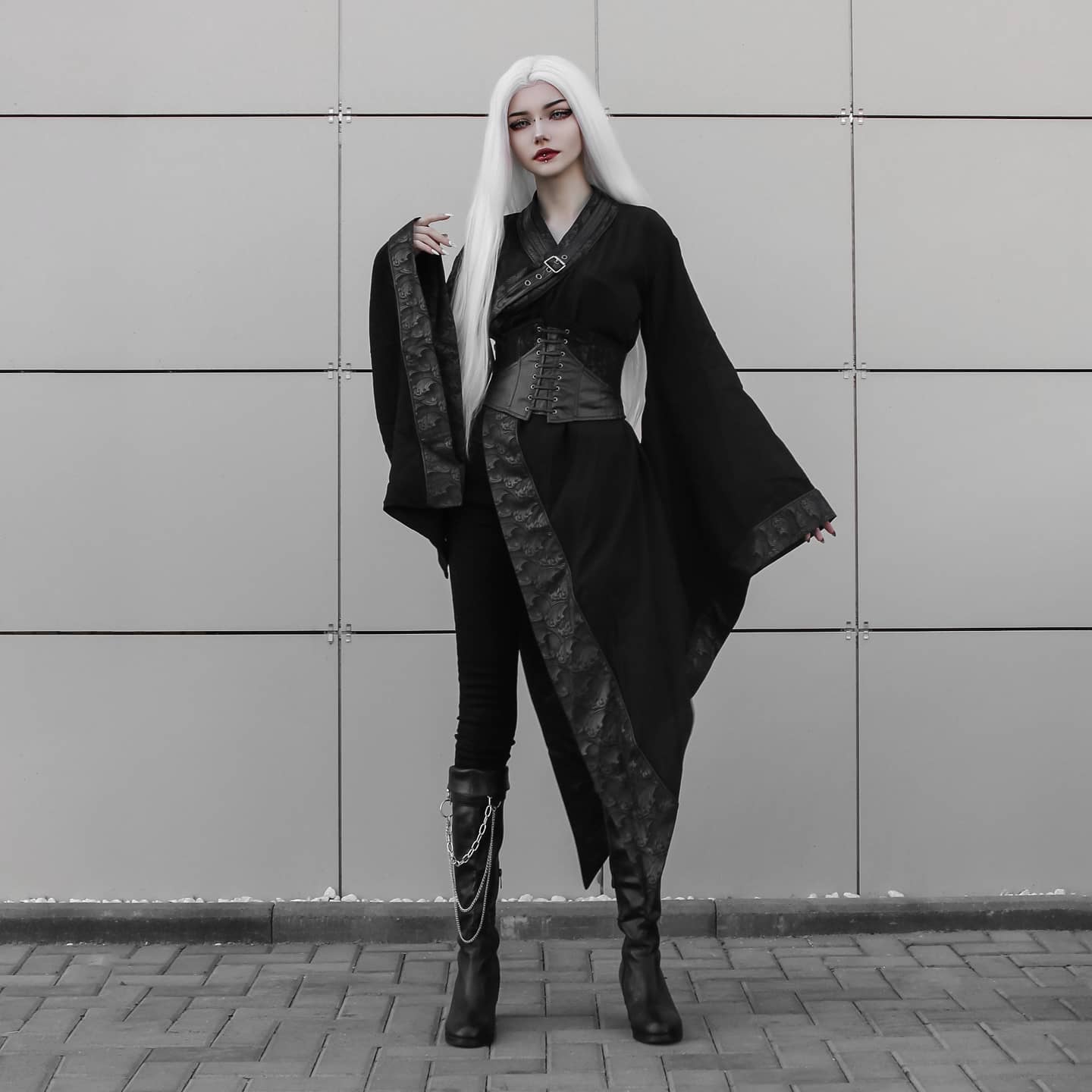 a fulllength shot of a model with long, straight white hair, wearing a dark gothic lolitainspired outfit. the outfit consists of a long black coat with kimonostyle sleeves and an asymmetrical hemline, paired with a black corset and tight black pants.  the model is also wearing tall black boots adorned with chains.  she stands against a grey tiled wall, creating a strong contrast with her dark attire. the overall style is dark, edgy, and fashionable, suggesting a gothic or cyberpunk aesthetic. the image has a cool, monochromatic feel.