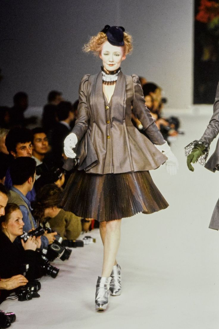 a model walks down the runway at a fashion show, wearing a brown jacket and a pleated skirt, both with an interesting and unusual design. she is also wearing silver boots, a black hat with a veil, and white gloves. the model is walking towards a group of photographers who are waiting to take her picture. there are also other models and audience members in the background. the overall scene is one of fashion and beauty, with a focus on the creative and unique designs of the clothes.