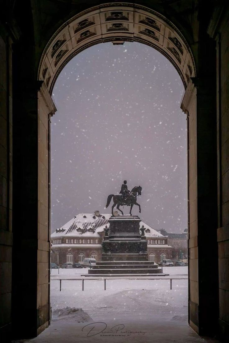 the image shows a view through an archway, possibly part of the brandenburg gate in berlin, germany.  it's a snowy winter day, with snow falling softly.  a large equestrian statue is visible in the distance, set upon a pedestal. behind the statue is a building with a snowcovered roof. the overall scene is cold and quiet, with a strong emphasis on the contrast between the dark archway and the snowy, lightfilled scene beyond. the image has a slightly vintage or moody feel.