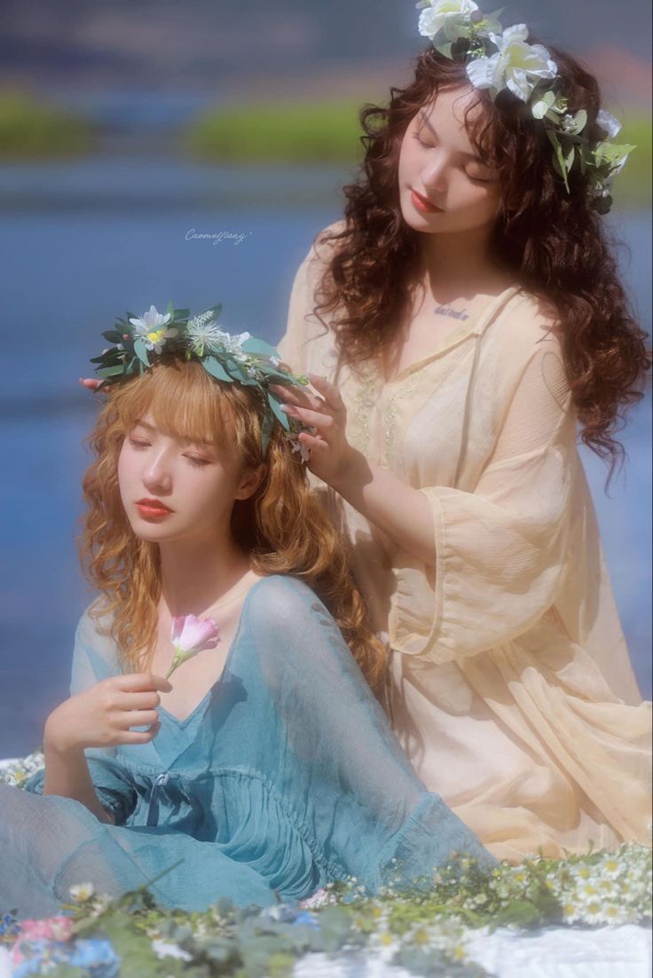 two young women wearing flower crowns embrace in a field of flowers. the woman in the blue dress is smiling sweetly, while the woman in the white dress looks lovingly at her. the image is soft and dreamy, with a whimsical and ethereal quality. it evokes feelings of love, friendship, and happiness.