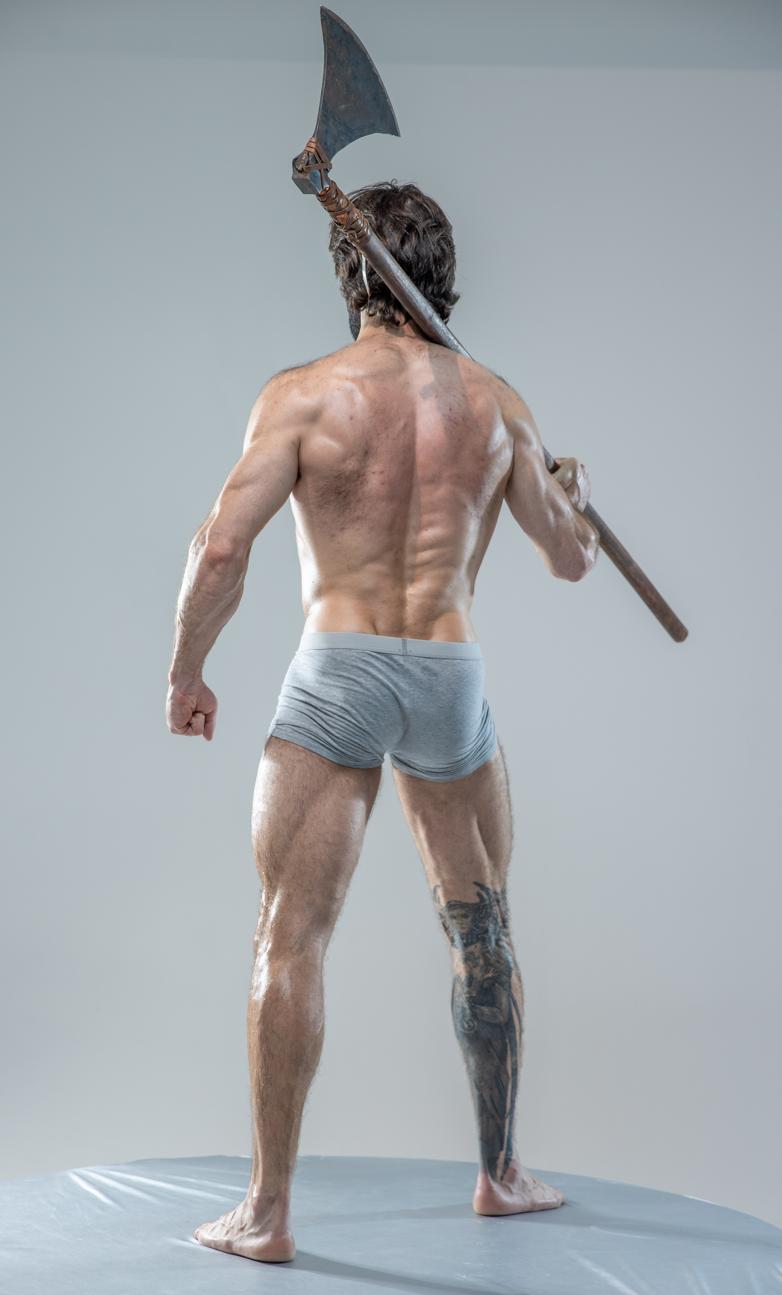 a muscular man with a beard and tattoos stands with his back to the camera, showcasing his toned physique. he holds a large, worn axe over his shoulder. he is wearing grey underwear and is positioned on a grey platform in a studio setting with soft, even lighting. the image evokes a sense of strength, power, and perhaps a historical warrior archetype.
