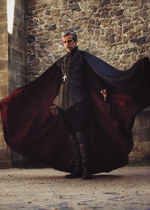 a full shot features peter capaldi in a dark, medievalstyle costume, standing outdoors against a brick wall. he is wearing a long, dark cape that is open, revealing a red lining. a cross hangs on a chain around his neck. the image is a portrait, capturing capaldi in character.
