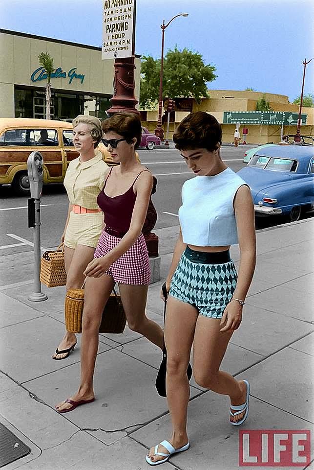 three women are walking on a sidewalk. they are wearing shorts and sandals. two of them are carrying straw bags. the woman in the middle is wearing sunglasses. there are cars parked on the street behind them. there is a building with a store sign visible in the background. a sign reads "no parking 7 am to 9 am, 4 pm to 6:15 pm, parking 1 hour, sam to 4 pm". a "life" logo is visible in the bottom right corner.