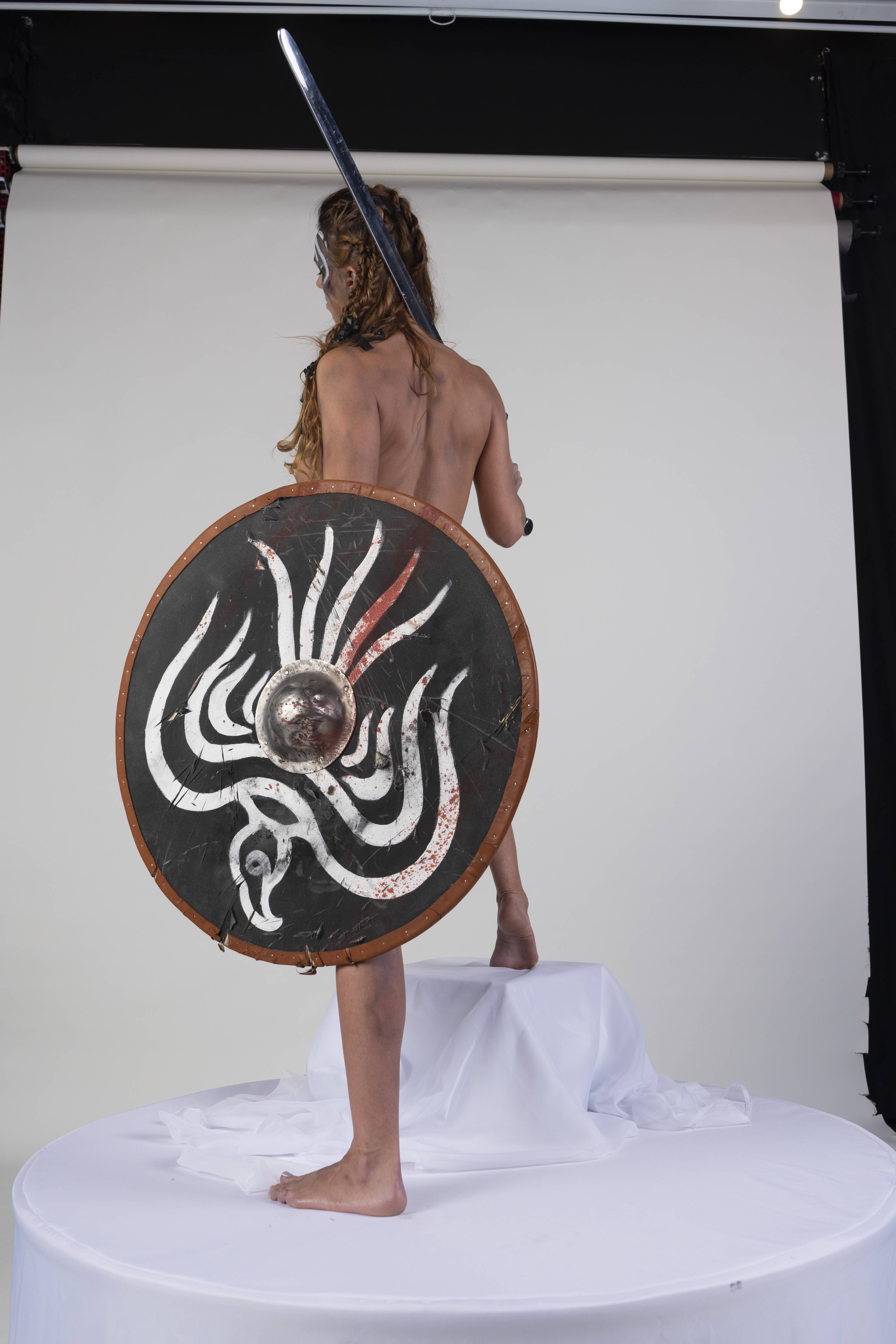 a shirtless person with braids stands with their back to the viewer in a studio setting. they hold a shield with a distinct design and a sword. the person is posing on a white platform covered in white fabric. the background is a plain white backdrop with studio equipment visible at the top.