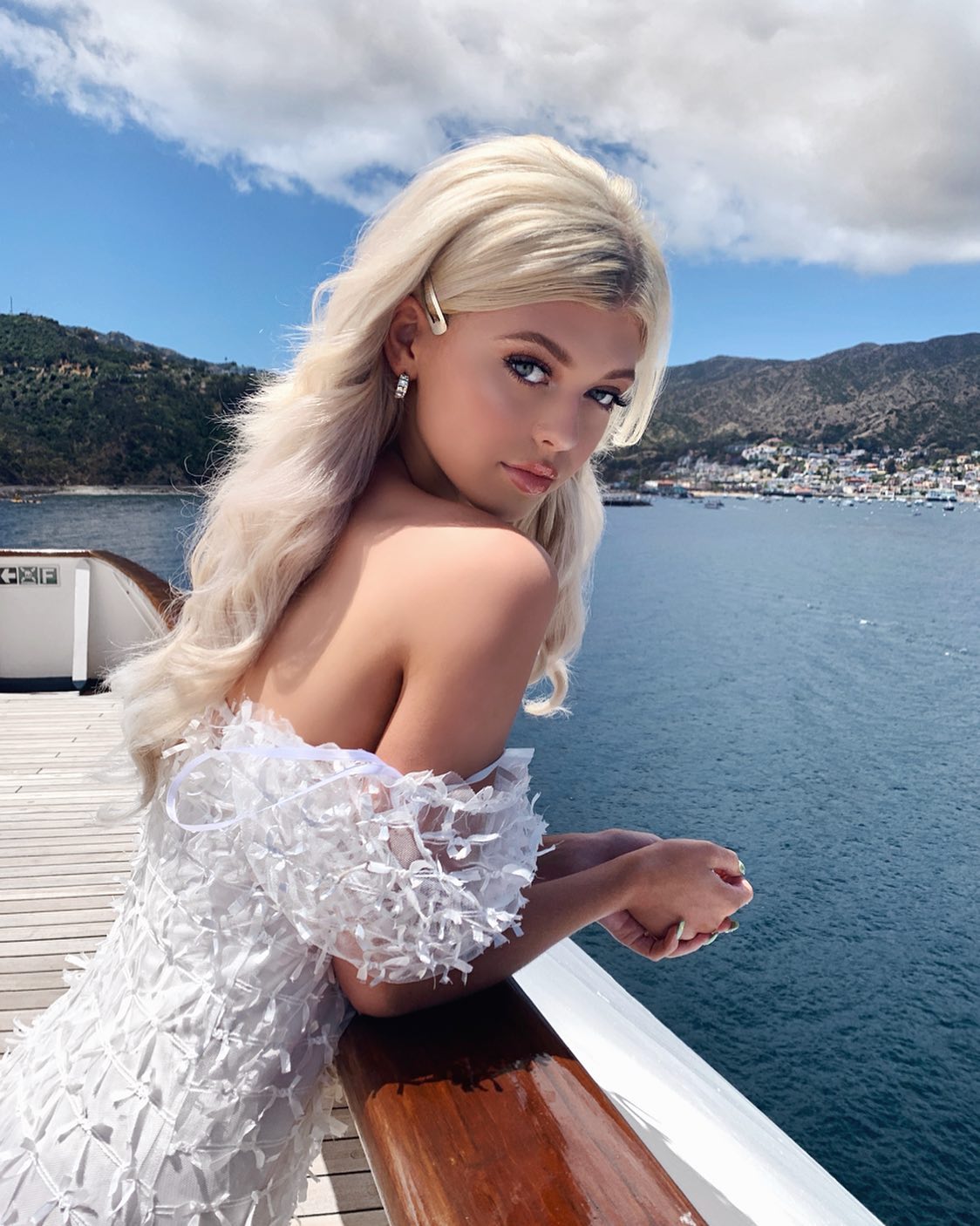 a young woman with blonde hair, wearing a white dress and hair clip, poses on the deck of a cruise ship with the ocean, mountains, coastline, blue sky, and clouds in the background.