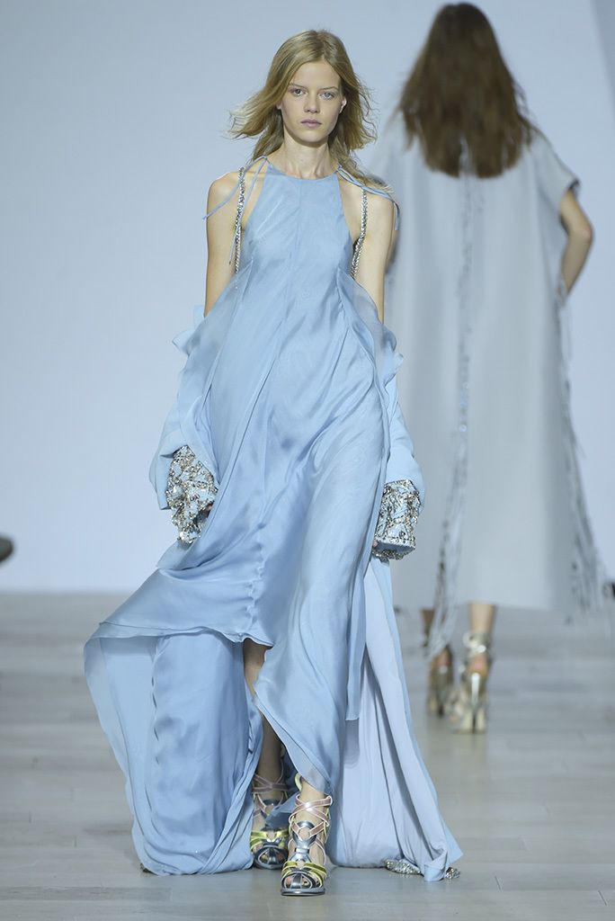 a fashion model in a light blue gown walks down a runway. her dress is flowing and elegant, and has a deep vneck, halter neckline, and a front slit. the dress is adorned with sparkly, silver jewelry and a long train. she is also wearing silver metallic high heel sandals. the model is looking down and to the right. there is another model in the background,  in a white  dress, walking towards the camera.